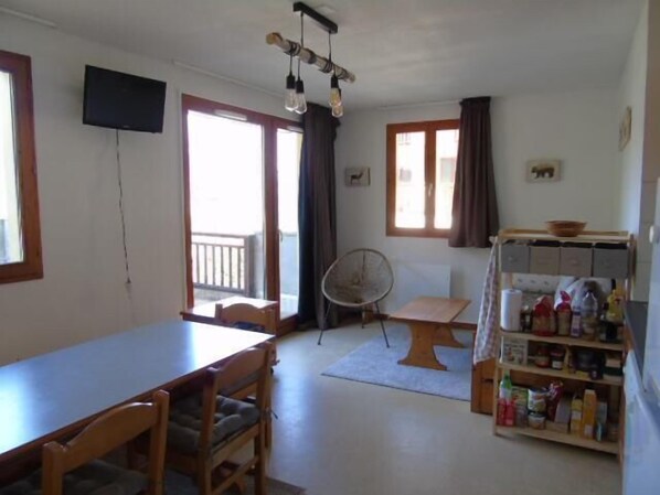 Une pièce comprenant une table à manger, des chaises, un ventilateur, une étagère avec des ustensiles de cuisine et un téléviseur fixé au mur.