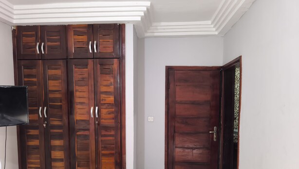 A room with a large wooden wardrobe, a closed door, and a television mounted on the wall. - SOTIDEK YOP (Abidjan)