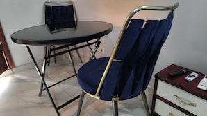 A room with a round table, a blue chair with a metal frame, and a small wooden side table with a drawer and a remote control. - SOTIDEK YOP (Abidjan)
