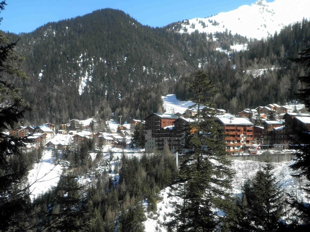 Estância de montanha coberta de neve com vários edifícios, incluindo um grande chalé com varandas, rodeada por florestas densas e encostas nevadas.
