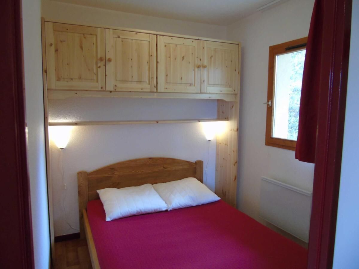 A single bed with a wooden headboard and a red bedspread, under a wooden cabinet with a window above.