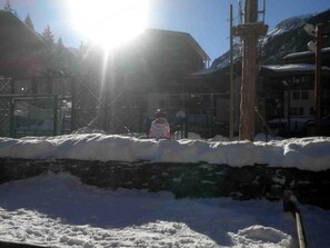 A snow-covered outdoor area with a stone wall, a person in winter clothing, and buildings in the background. - Ground floor, view mountain, tv, ski locker, 28m², Valfréjus (Valfréjus)