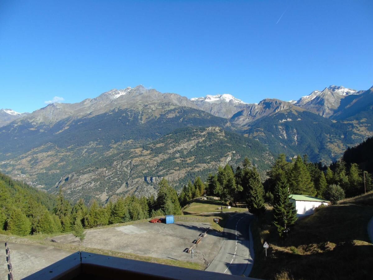 A mountainous landscape with a clear sky. There are trees and a small building visible.