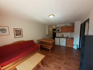 A compact living space with a red sofa, wooden dining table, kitchenette with cabinets, and a small white trash bin.