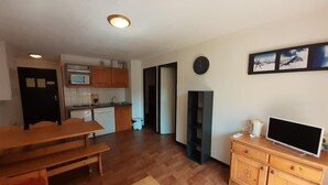 Um espaço compacto com mesa de jantar de madeira, TV sobre um móvel de madeira e cozinha com micro-ondas e armários.