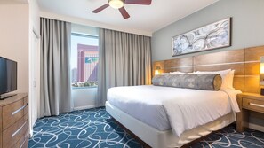 Une chambre d’hôtel avec un grand lit, une télévision, un ventilateur de plafond, une fenêtre avec des rideaux, une œuvre d’art accrochée au mur et une moquette à motifs.