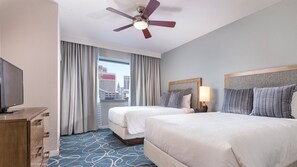 Une chambre d’hôtel avec deux lits, un ventilateur de plafond, une télévision et une vue sur la ville par la fenêtre.