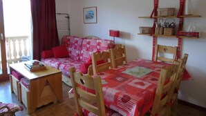 A cozy room with a red sofa, wooden dining table, and chairs. There is a small table with a book and a red lamp. A shelf with baskets and a framed picture is on the wall. - 5th floor, view mountain, balcony, tv, ski locker, 34m², Valfréjus (Valfréjus)