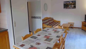 Uma área de refeições com uma mesa coberta por uma toalha estampada, um sofá, uma pequena mesa de madeira e um relógio fixado na parede.