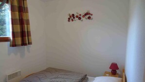 Quarto pequeno e simples com uma cama, uma decoração na parede, uma janela com cortinas e um abajur.
