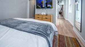 A modern bedroom with a large bed, a flat-screen TV mounted on the wall, a wooden dresser, and a hallway leading to another room. - OTR, 1blk to TQL, Cali King Bed, Sleeps 4, Free Light Rail, 58in TVs, Wifi (Cincinnati)