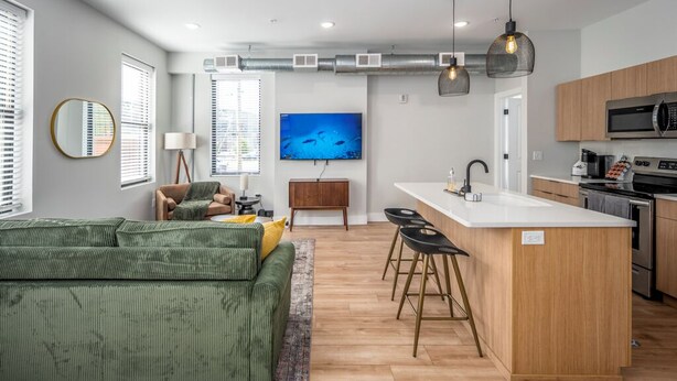 A modern living room with a green sofa, a small wooden side table, and a TV mounted on the wall. - OTR, 1blk to TQL, Cali King Bed, Sleeps 4, Free Light Rail, 58in TVs, Wifi (Cincinnati)