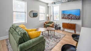A living room with a green sofa, yellow cushions, a coffee table, a side table, a lamp, a TV, and a wooden cabinet. - OTR, 1blk to TQL, Cali King Bed, Sleeps 4, Free Light Rail, 58in TVs, Wifi (Cincinnati)