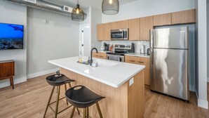 A modern kitchen with a white island, stainless steel appliances, wooden cabinets, and two black bar stools. - OTR, 1blk to TQL, Cali King Bed, Sleeps 4, Free Light Rail, 58in TVs, Wifi (Cincinnati)