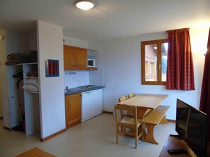A compact kitchen with a microwave, stove, and wooden cabinets. There is a dining table with chairs and a TV on a stand.