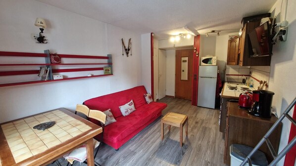 A compact living space with a red sofa, a small wooden table, a kitchenette, and a red-framed mirror.