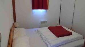 A small, single-bed room with a wooden headboard, a red blanket, and a window with a red curtain. - 4th floor, view valley, balcony, tv, ski locker, 26m², Valfréjus (Valfréjus)