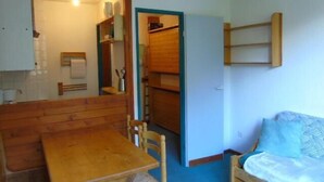 Um quarto pequeno e acolhedor com uma mesa de madeira e cadeiras, um banco, uma cama com travesseiro estampado e uma prateleira de madeira com diversos objetos.