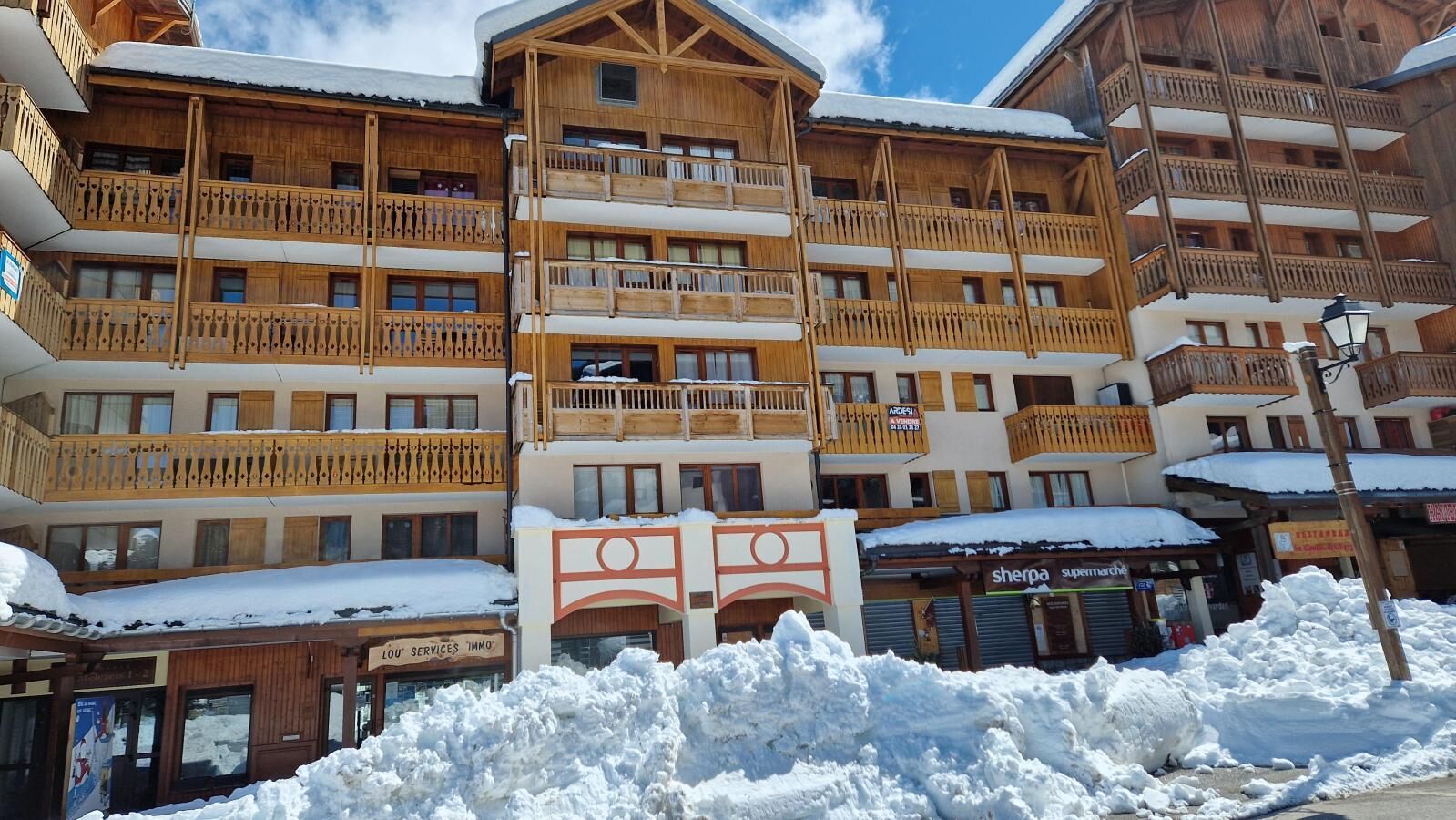 A multi-story wooden building with balconies, a sign for 'Sherpa Supermarché', and snow on the ground.