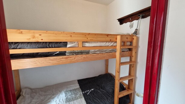 Una cama litera con estructura de madera y una escalera, una pared blanca y un marco de puerta rojo.