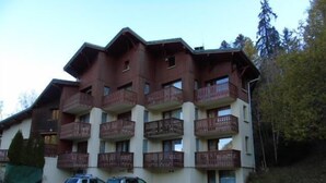 Un edificio a più piani con balconi, un'area di parcheggio con auto e uno sfondo naturale.