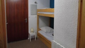 Una stanza piccola e accogliente con una porta in legno, un letto singolo con materasso e cuscini, un tavolino bianco con una lampada e un termosifone bianco.