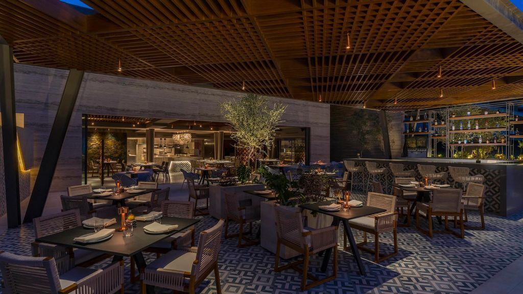 Un área de comedor al aire libre con mesas y sillas, un bar con estantes, y un árbol grande en el centro.