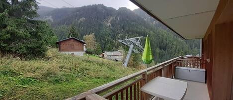 A balcony with a table and a green umbrella, overlooking a forested valley with a mountain in the background.