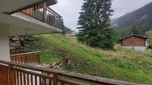 Un balcón con barandilla de madera que da a una zona montañosa con vegetación y un edificio pequeño a lo lejos.