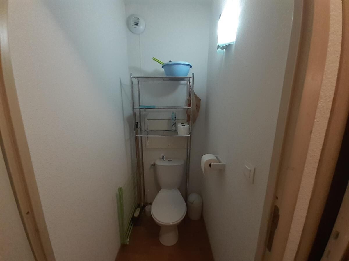 Ett litet, smalt badrum med en toalett, en hylla med en skål och en toalettpappersrulle samt en papperskorg.