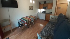 Un espacio habitable compacto con una mesa de comedor, un sofá, un televisor montado en la pared y una kitchenette al fondo.