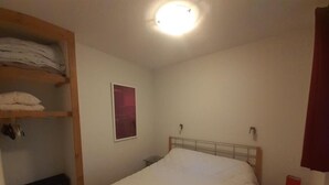 Una habitación pequeña y sencilla con una cama, un estante con ropa doblada, una mesita y una lámpara en la pared.