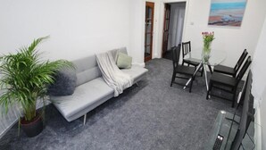 A living room with a grey sofa, a coffee table, and a dining table with chairs. There is a potted plant and a painting on the wall.