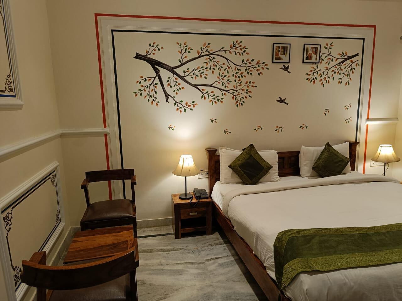 A hotel room with a bed, two bedside tables with lamps, a chair, and a decorative wall with a tree and bird mural.
