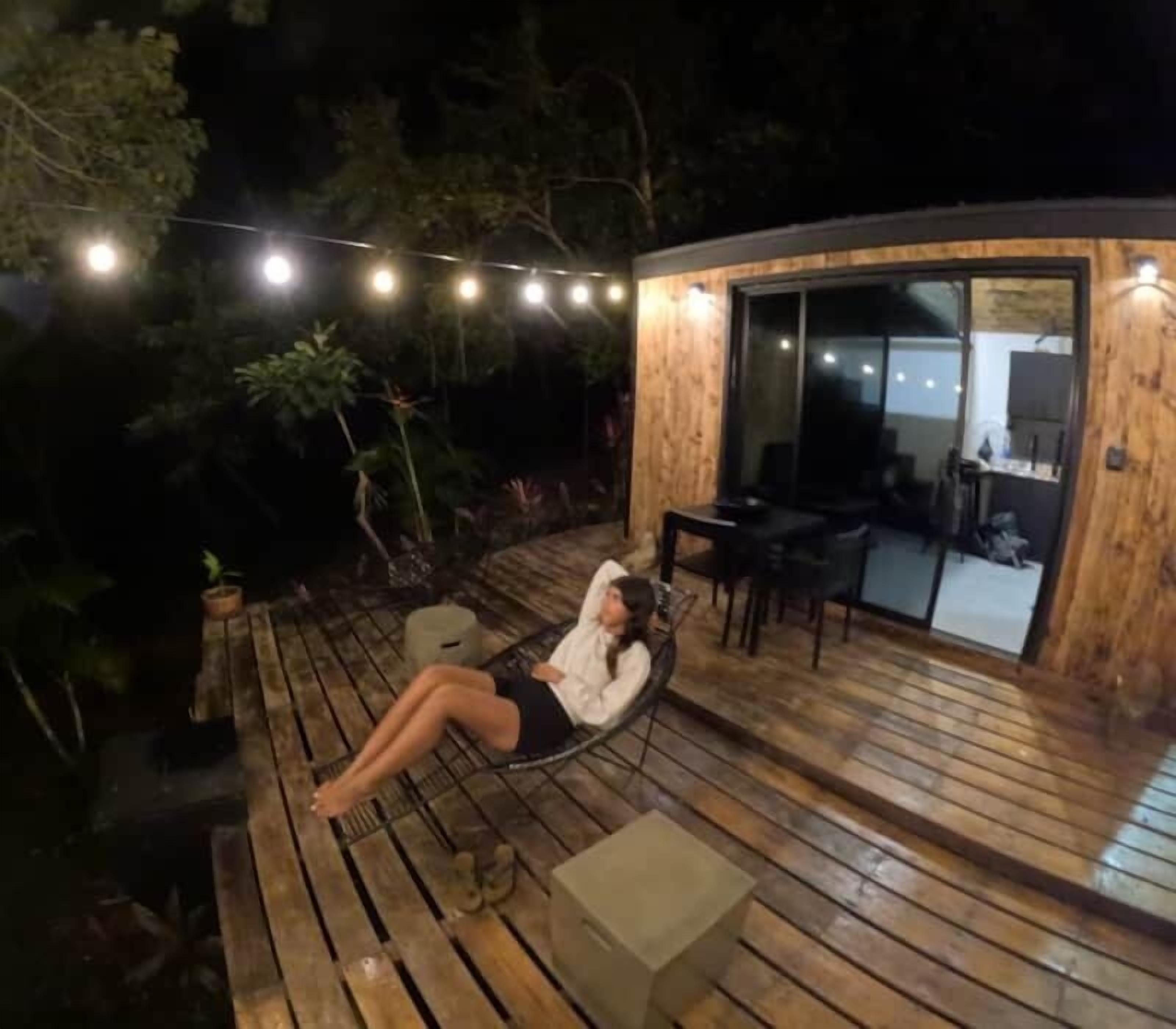 A person lying on a wooden deck with string lights overhead, a glass door leading to an interior space, and potted plants around.