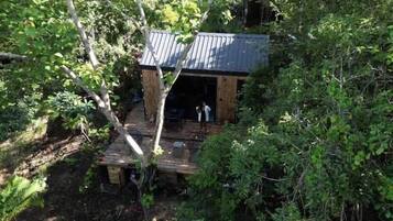 A rustic cabin with a wooden deck and a metal roof, surrounded by dense foliage.