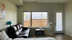 A bedroom with a black leather sofa, a small wooden table, a standing fan, and a window with a view of a wooden fence and buildings.