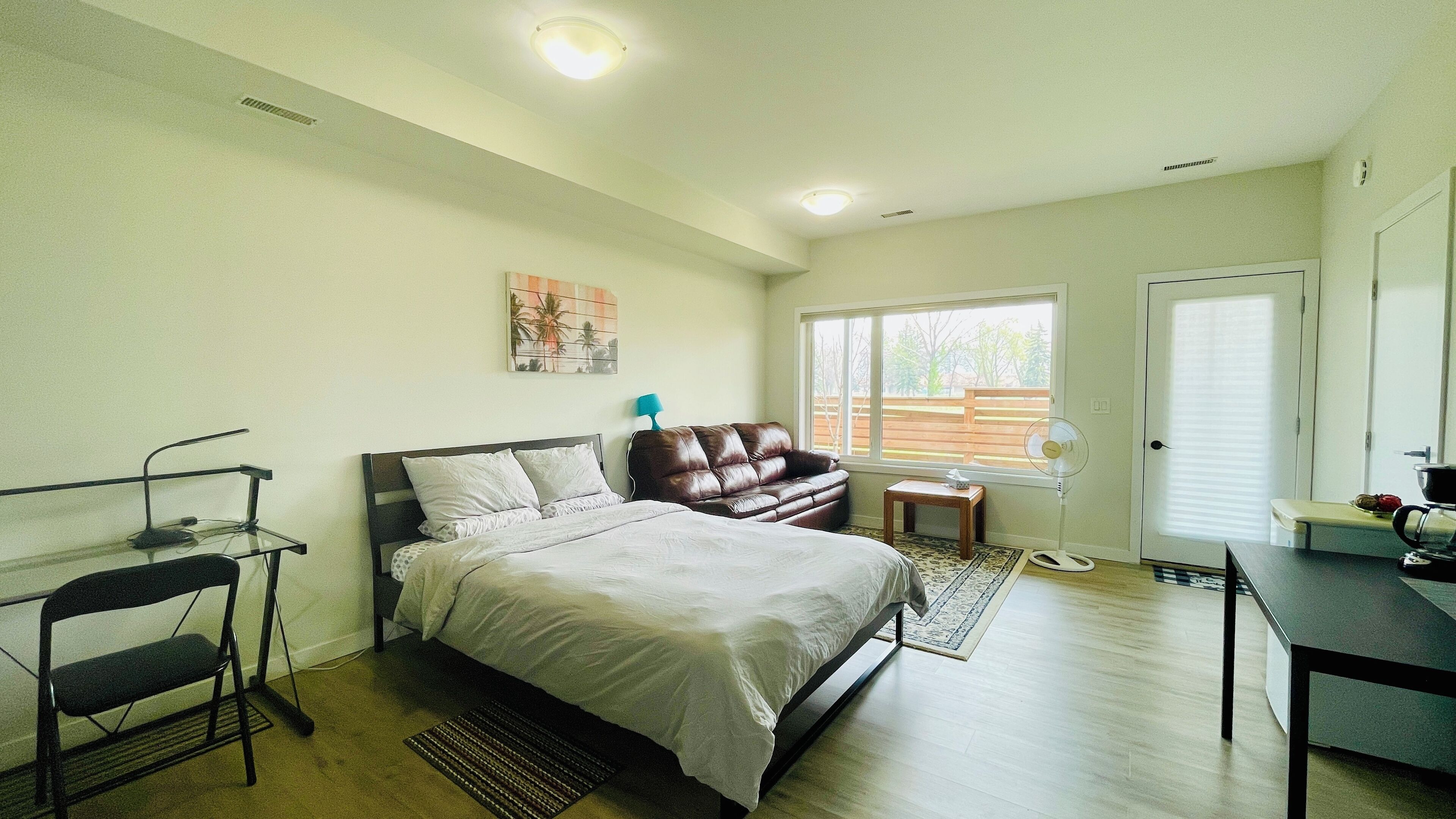 Un dormitorio con cama, escritorio, silla, ventilador y ventana con vistas al exterior.