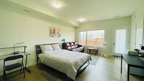 A bedroom with a bed, a desk, a chair, a standing fan, and a window with a view of the outdoors.