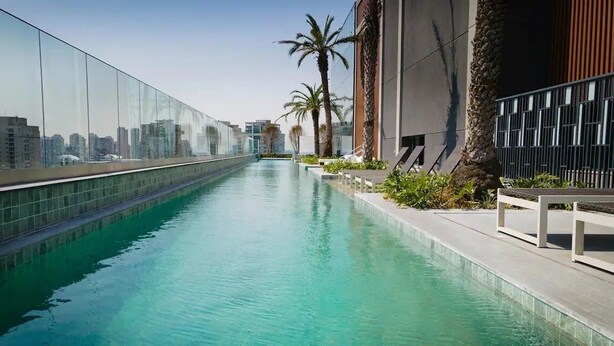 A modern rooftop pool with clear blue water, surrounded by glass walls and palm trees, offering a view of the city skyline. - Get a Flat 1905 - Brooklin (São Paulo)