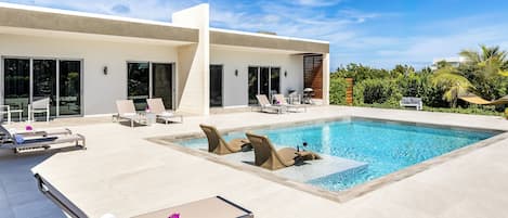Un área de piscina moderna con sillones y una mesa, rodeada por un edificio con amplias ventanas y una piscina.