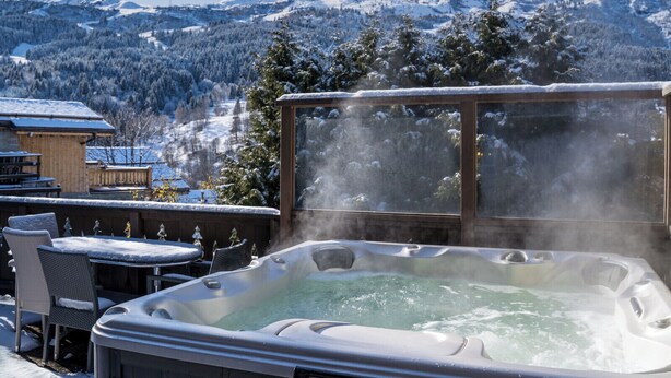 Un jacuzzi con vistas a montañas nevadas y una terraza de madera con mobiliario exterior.
