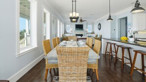 A dining area with a long table, wicker chairs, and a kitchen in the background.
