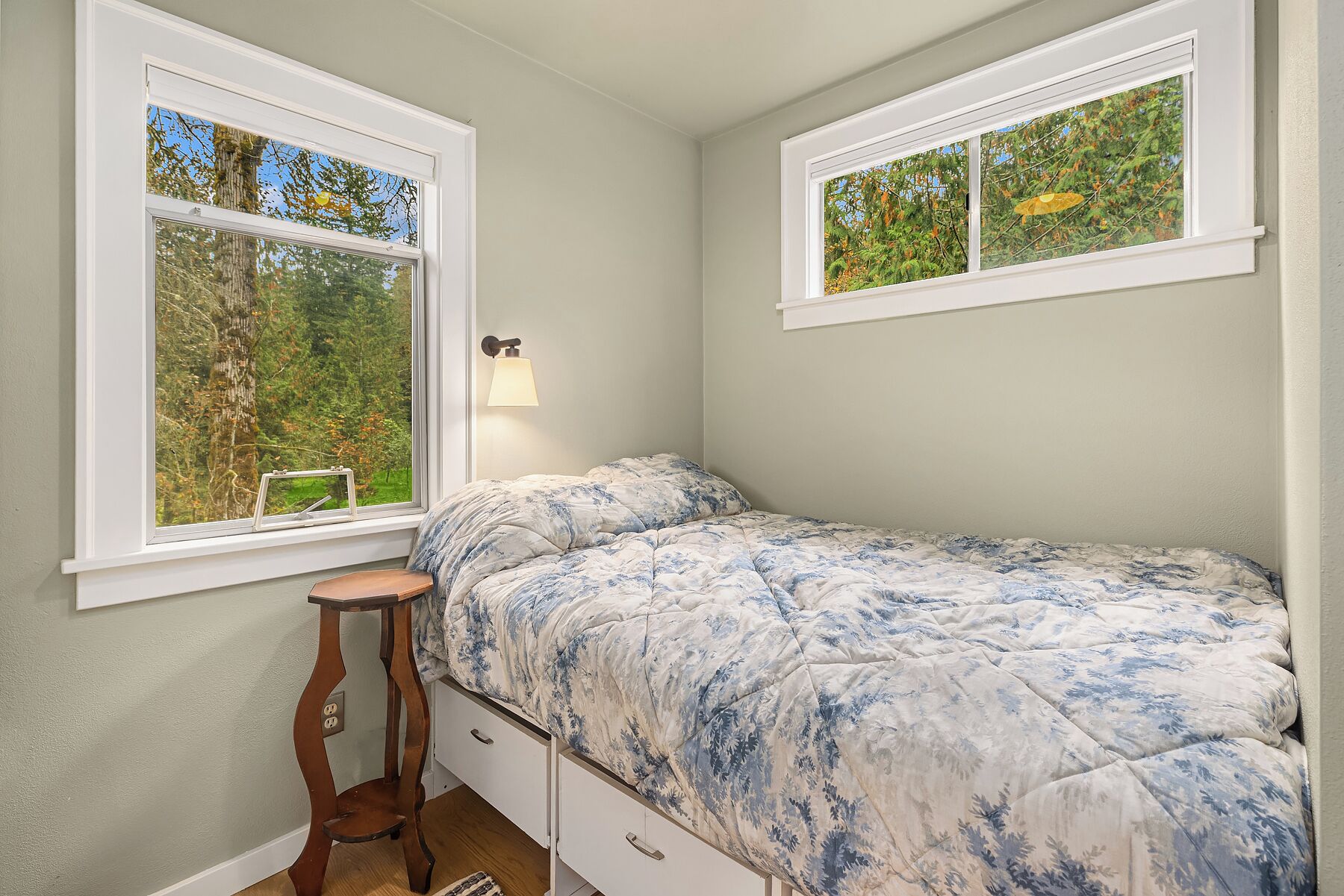 A bedroom with a single bed, two windows, a bedside table, and a lamp.