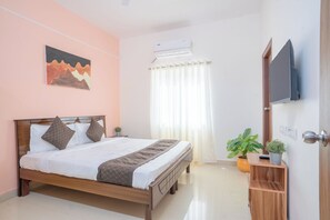 Un lit bien fait, avec un cadre en bois, un téléviseur fixé au mur, une plante en pot et une fenêtre avec des rideaux.