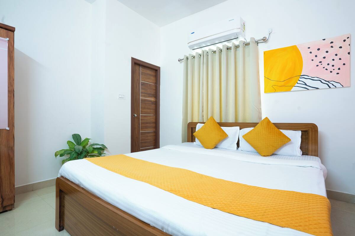Una cama bien hecha con estructura de madera, ropa blanca de cama y dos almohadas amarillas. A la izquierda de la cama hay una planta en maceta, y arriba de ella un aire acondicionado instalado en la pared.