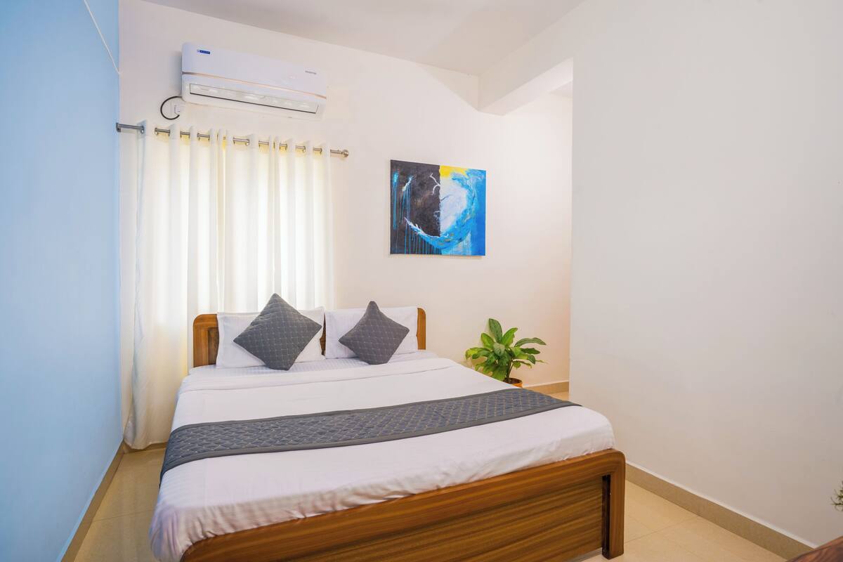 Un dormitorio con cama, cortinas, aire acondicionado de pared, un cuadro y una planta en maceta.