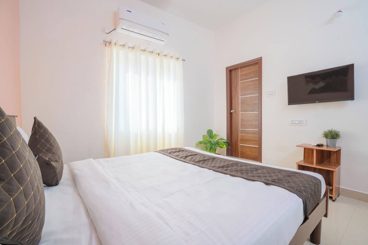 Un dormitorio con cama, cortinas, aire acondicionado, una puerta de madera, un televisor y una planta pequeña.