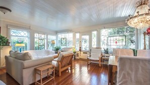 A spacious living room with a wooden floor, white furniture, and large windows offering views of greenery. - Vintage Cottage steps from Venice Beach (Los Angeles)