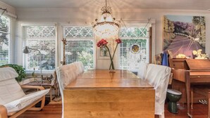 A dining area with a wooden table, white upholstered chairs, a chandelier, and a painting on the wall. - Vintage Cottage steps from Venice Beach (Los Angeles)